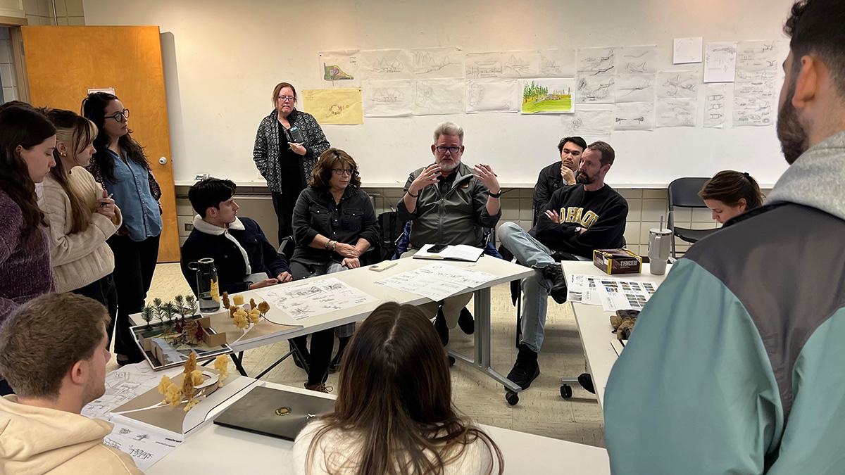 Photo of students and faculty sitting around tables with drawings in the background.